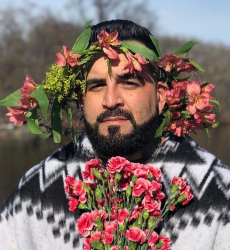 Queer nonbinary Latinx author Mark Oshiro has dark hair, facial hair, and medium skin tone. They have flowers encircling their head in a crown and are holding flowers.