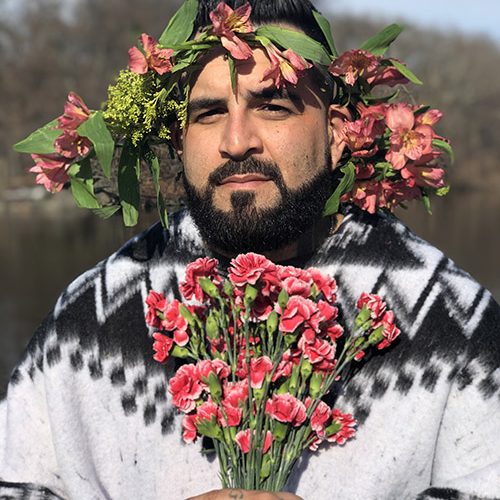 Man with medium skin tone, and a dark beard mustache wearing a wreath of pink flowers and green leaves on his head, holding a bouquet of pink carnations wearing a black and white sweater with geometric design