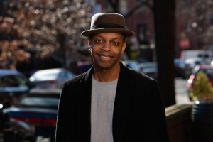 Robert Jones, Jr. smiling on a street. He is wearing a brown fedora and a black jacket over a gray shirt.