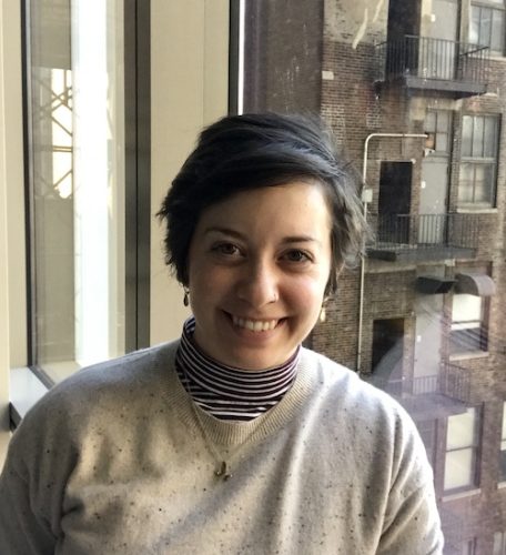 Anna Membrino, is a queer author with short dark hair, light skin tone. She is wearing small earrings and a turtleneck and light-colored sweater. She is smiling directly at the camera.