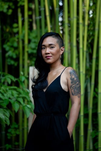 Indonesian-Canadian Queer poet Cynthia Oka, a woman with dark long hair, shaved on one side, brown eyes, and medium skin tone, with a large tattoo on her shoulder and down her upper arm. Green bamboo trees in the background. She is wearing a black dress with spaghetti straps and her hands are in her pockets.
