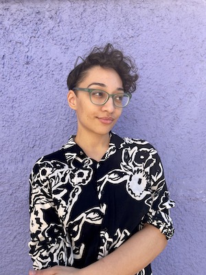 Woman with medium skin tone and short curly hair wearing a patterned black and white short sleeved shirt with collar and glasses
