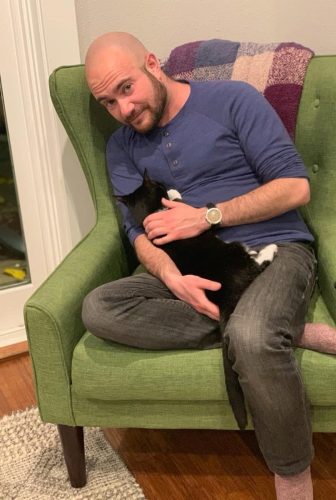 An illustration of transmasculine queer author Kyle Lukoff, a man with a brown beard, brown eyes, and light skin tone. He is holding a black and white cat in his lap, and is sitting in a green chair with a plaid blanked draped over the back
