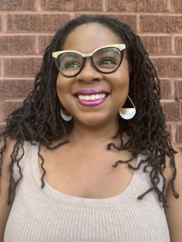 Woman with dark skin tones and locs wearing gold and black eyeglasses, earrings and a beige tank top, with a brick wall in background