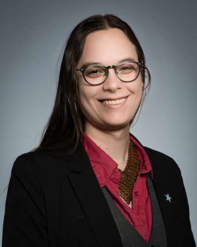 Tobi Hill-Meyer is an Indigenous Chicana trans woman. She has light toned skin and long dark hair parted in the middle. She is wearing a dark red collared shirt and a dark jacket. She is smiling at the camera