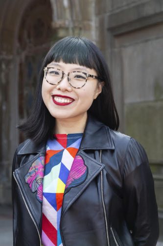 Chinese-American queer writer, illustrator, and cartoonist Yao Xiao, a woman with dark straight hair with bangs, light skin tone, and glasses. She is wearing a black jacket with lapels and a shirt with multi-colored geometric patterns.