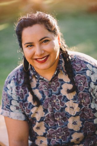 Image of Sonora Reyes, queer author light-medium skin tone dark hair in two braids, big gold hoop earrings and dark red lipstick smiling and wearing a flowered blue, purple and light pink short sleeved collared shirt