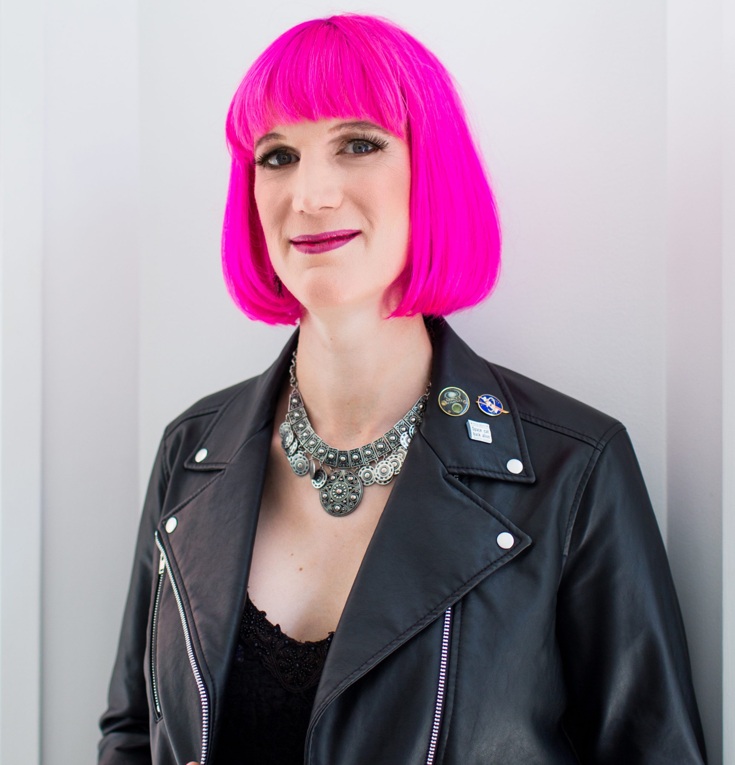 Charlie Jane Anders is pictured in front of a white background. They have light-toned skin and are wearing a black sleeveless dress. They have chin-length hair and bangs colored bright pink.