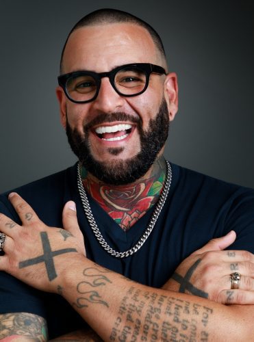 A headshot of Mark Oshiro. They have light brown skin and their arms are crossed over their chest. Their tattoos on their arms, hands, and chest are visible. They are wearing dark-framed glasses and smiling wide.