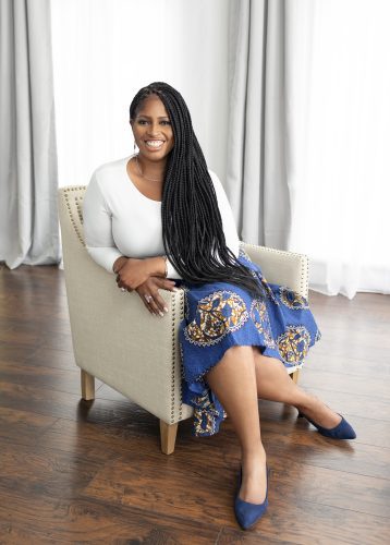 Mecca Jamilah Sullivan sits on white chair wearing long braids and a blue African print skirt.