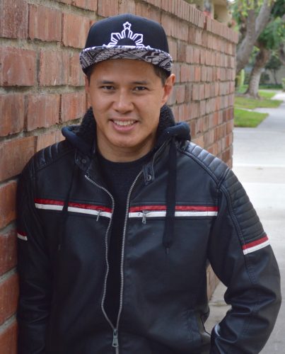 A Filipino American man smiles in front of a brick wall. He wears a cap and a leather jacket with red and white striped details.