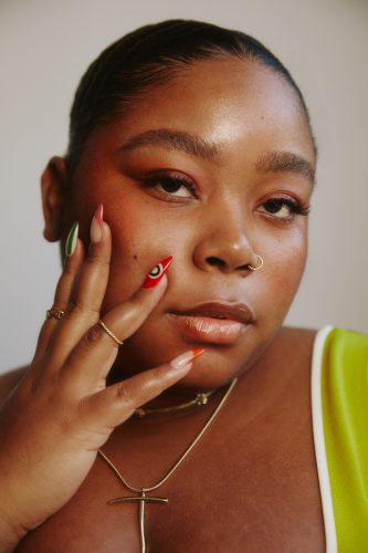 Portrait of Tembe Denton-Hurst against a grayish beige background, with her hand on her face. She is wearing a gold necklace, rings, and her green and red nail art designs.