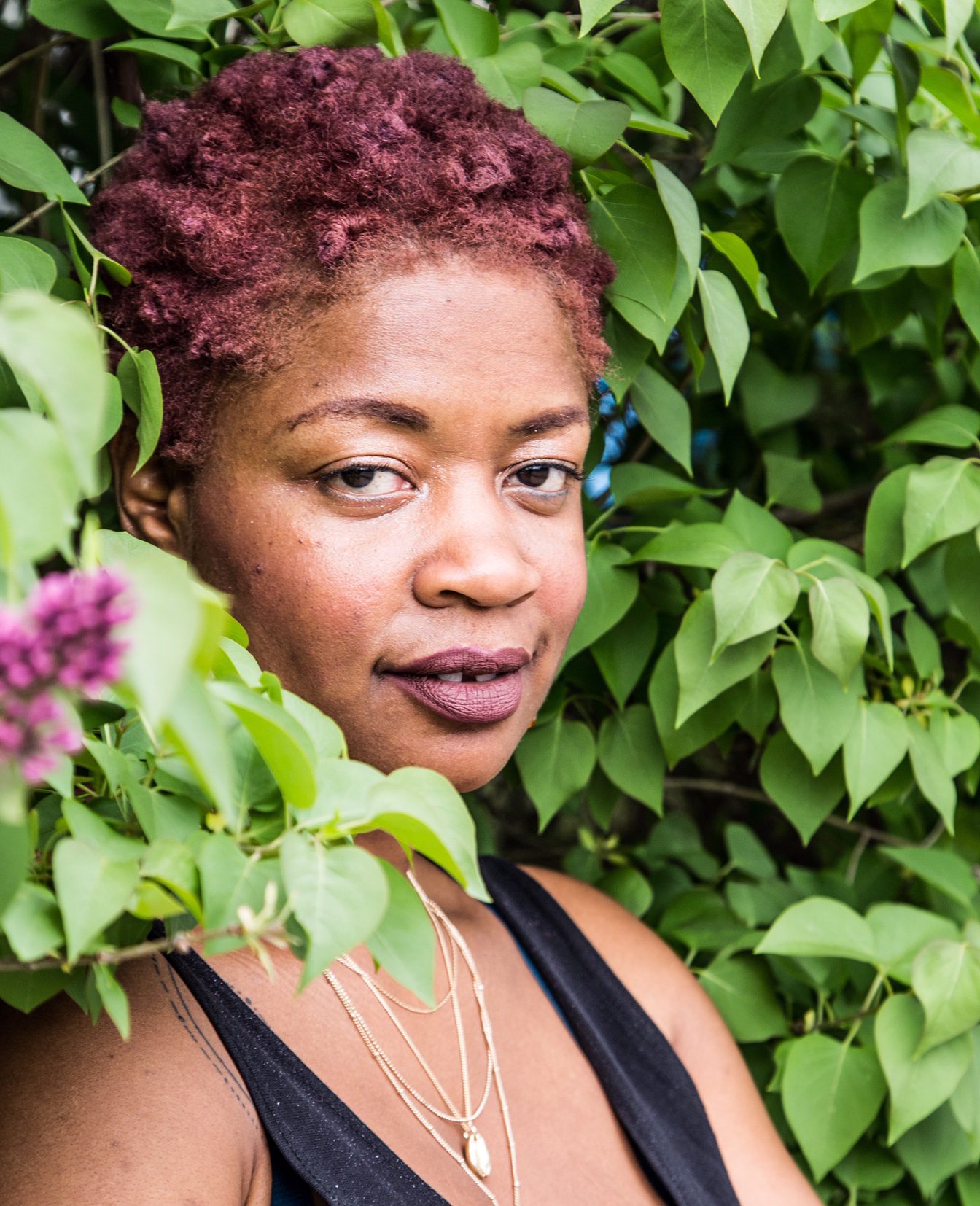 Black woman with magenta curly afro leaning into a background of foliage with a slight smile.