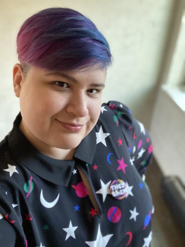 Image of Christine, who has blue and purple hair and is wearing a black dress with colorful stars, moons, and planets on it. They are also wearing a pronoun pin that says "they/them"
