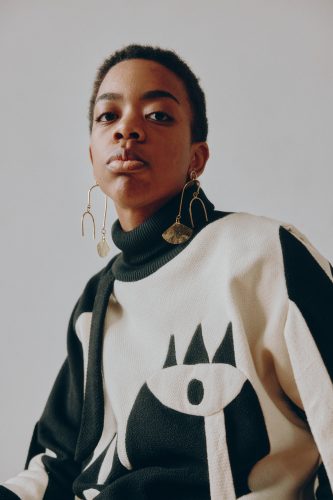 Dale Walls, a Black American person with a short haircut looks slightly downward towards the viewer. They are wearing a black and white sweater that features an eye on the design and dangling gold earrings.