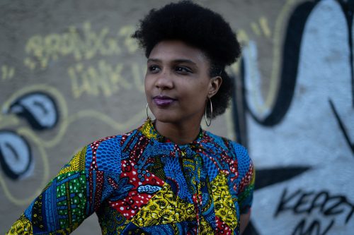 The author is posing in front of a graffiti art piece outside in her Flatbush neighborhood. She is wearing a brightly colored Afro-inspired shirt dress from Culture Chest. Her face is gazing in the distance. Her hair is in a half-afro and she wears hoop earrings. She is Black.